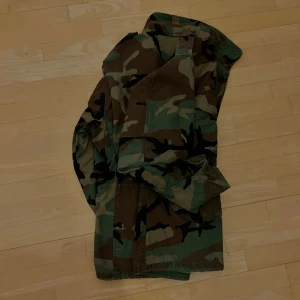 Camouflage militärjacka - Eftertraktad och trendig kamouflage militär jacka i gott skick.  Modell: M-65, klassisk militärjacka, började tillverkas 1965 för US Army Str: M  *Som storleksreferens är jag 1.94 lång, jackan är ngt för kort för mig, helt enkelt en normal M med plats för en extra tjock tröja under.  Dragkedja och knappar samt två fickor framtill plus smart innerficka, huva smart gömd i kragen.  Seriös slitstark kvalitet, fodrad med dubbelt tyg. 