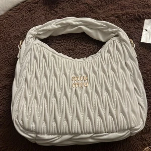 Vit axelväska  - Säljer en elegant vit axelväska från M med quiltad design och ett stilrent handtag. Väskan har en gyllene logotyp på framsidan som ger en lyxig touch. Perfekt för att lyfta din outfit till nästa nivå.