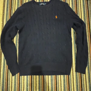 Mörkblå stickad tröja från Ralph Lauren - Säljer en snygg mörkblå stickad tröja från Ralph Lauren med kabelstickat mönster. Tröjan har långa ärmar och en liten orange logotyp på bröstet lite solblekt längst up på kragen. Perfekt för kyligare dagar.