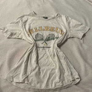 Vit t-shirt från Gina Tricot - Säljer en vit t-shirt från Gina Tricot med tryck av två tennisracketar och texten 'Wellbeing Tennis Court Tournament 1998'. Perfekt för en sportig look eller casual vardagsstil.