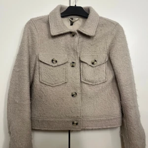 Beige jacka från H&M - Säljer en snygg beige jacka från H&M med knappar framtill och två bröstfickor. Jackan har en klassisk krage och är tillverkad i ett mjukt material som ger en stilren look. Perfekt för kyligare dagar!