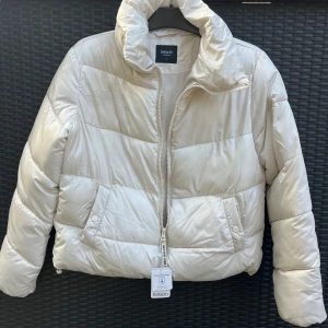 Vit pufferjacka från DeFacto - Snygg vit pufferjacka i nyskick från DeFacto med dragkedja framtill. Jackan har en hög krage och två fickor på sidorna. Perfekt för kyliga dagar när du vill hålla dig varm och stilren. 
