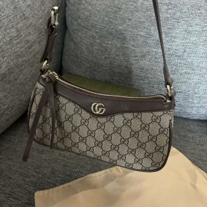Elegant axelväska med GG-mönster - Snygg axelväska helt oanvänd från Gucci med det ikoniska GG-mönstret. Väskan har en dragkedja och justerbar axelrem. Finns i olika nyanser av beige och grått med stilrena detaljer i skinn. Finns även box!