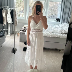 Vit sommarklänning med spetsdetaljer - Säljer en vacker vit klänning med spetsdetaljer och v-ringad design. Klänningen har smala axelband och en öppen rygg med knytning. Perfekt för sommarens varma dagar. På sista bilden visas dock en defekt vilket är att ena axelbandets fästning på ryggen har lossnat men vi knöt ihop den. Annars går det enkelt att sy ihop. Det är också en del av broderiet som har lossnat någon centimeter på framsidan vilket syns på bild 1 och 2 men som ni ser är det inte särskilt märkbart och kan också sys ihop.