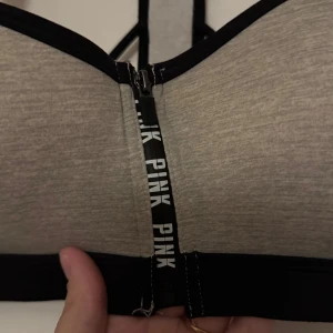 Grå sporttopp från Victoria's Secret PINK - Säljer en grå sporttopp från Victoria's Secret PINK med svarta detaljer och dragkedja framtill. Toppen har en snygg ryggdesign med rosa inslag och är perfekt för träning. Är en lätt push up! Strl M/ 75/80B 