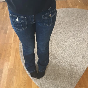 Mörkblå jeans med fickdetaljer - Snygga mörkblå jeans med framträdande vita sömmar och fickor med knappar framtill. Byxorna har en bootcut-stil som ger en klassisk look. Perfekta för en avslappnad stil.