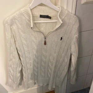Vit stickad tröja från Polo Ralph Lauren - Säljer en stilren vit stickad tröja från Polo Ralph Lauren med dragkedja och broderad logga på bröstet. Tröjan har en klassisk kabelstickad design och långa ärmar. Perfekt för kyligare dagar!