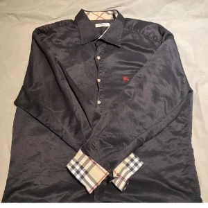 Svart skjorta från Burberry - Snygg svart skjorta från Burberry med klassiskt rutigt mönster på insidan av kragen och ärmsluten. Skjortan har knappar framtill och en liten broderad logga på bröstet. Perfekt för en stilren look. Storlek L men passar M. Pris kan diskuteras vid affär✌🏽