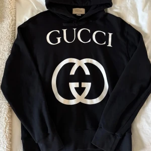 Svart hoodie från Gucci - Säljer en stilren svart hoodie från Gucci med det ikoniska logotyptrycket i vitt på framsidan. Tillverkad i Italien av 100% bomull, vilket gör den både bekväm och hållbar. Perfekt för en avslappnad och trendig look. Använd en gång.