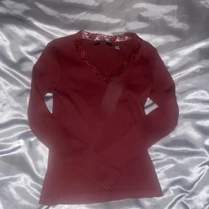 Vinröd ribbad topp  - Snygg vinröd ribbad topp från Vero Moda med spetsdetaljer vid v-ringningen. Den har långa ärmar och är perfekt för en stilren look. Passar bra till både jeans och kjol. Perfekt för höst och vinter!
