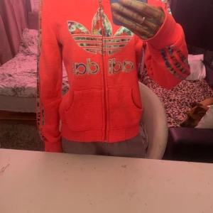 Rosa hoodie från Adidas - Säljer en snygg rosa hoodie från Adidas med blommigt mönster på loggan och längs ärmarna. Tröjan har dragkedja och huva med blommigt foder. 🌸