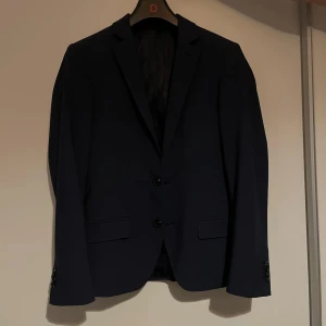 Mörkblå kostym från Dressmann - Snygg mörkblå kostym från Dressmann. Kavajen har två knappar och klassisk design. Perfekt för studenten! Säljer pga den är för liten, använd vid ett tillfälle vilken var studenten och är kemtvättas efter de! Kavajen är i storlek 44S och byxorna är i storlek 31-30.