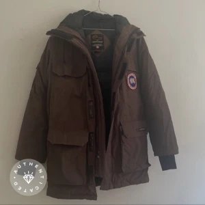 Brun parkajacka från Canada Goose - Säljer en brun parkajacka från Canada Goose med pälsdetaljer på huvan. Jackan har flera praktiska fickor och en dragkedja framtill. Perfekt för kalla vinterdagar. Använd men har väldigt mycket mer att ge! En liten skråma på jacka. Inget som stör något större. Se bild. 