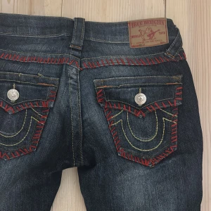 Mörkblå jeans med röda detaljer från True Religion - Snygga mörkblå jeans från True Religion med unika röda sömmar och stora knappar på bakfickorna.  Lågmidjade och utsvängda, innerben: 82, midja 34 rakt över, grenhöjd 18. Är 165 med långa ben som referens, går helst inte ner i pris men vid ett SNABBT köp så kan jag bjussa lite☺️