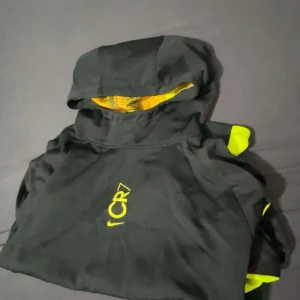 Nike CR7 - Säljer en svart hoodie från Nike med CR7-logga i gult. Hoodien har en praktisk ficka framtill och är tillverkad i Dri-FIT-material för optimal komfort. Den har långa ärmar och en huva med ett coolt mönster i gult och rött på insidan.
