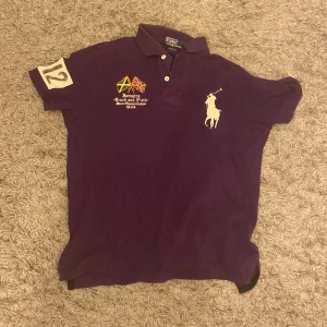 Lila pikétröja från Ralph Lauren - Säljer en snygg lila pikétröja från Ralph Lauren med broderade detaljer och nummer 12 på ärmen. Tröjan har en klassisk krage och korta ärmar. Perfekt för en avslappnad stil med en touch av elegans.