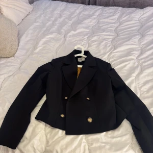 Svart kavaj från Gold Carino - Snygg svart kavaj från Gold Carino med dubbelknäppning och guldknappar. Perfekt för en stilren look. Kavajen har en klassisk krage och är långärmad. Passar bra till både formella och avslappnade tillfällen.