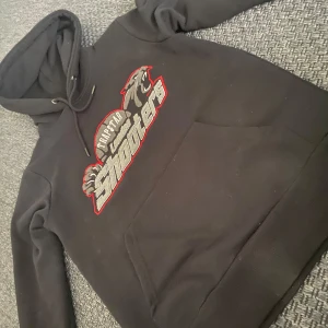 Svart hoodie från Trapstar - Säljer en svart hoodie från Trapstar med tryck av 'London Shooters' i grått och rött. Hoodien har en klassisk design med dragsko i huvan och en stor magficka framtill. Perfekt för en casual look.