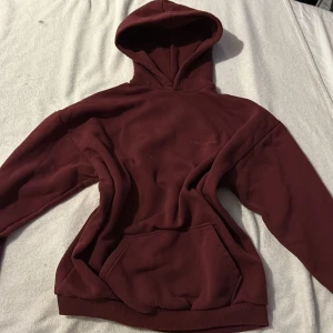 Vinröd hoodie från Boohoo - Säljer en vinröd hoodie från Boohoo med en mysig känsla. Den har en klassisk design med en stor ficka framtill och en huva. Perfekt för kyliga dagar och avslappnade stunder. Märket DSGN STUDIO är diskret tryckt på bröstet.