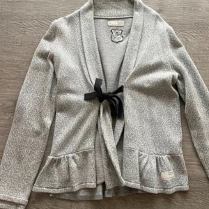 Grå mönstrad kofta från Odd Molly - Säljer en snygg grå kofta från Odd Molly med ett diskret mönster och volangdetaljer vid nederkanten. Koftan har långa ärmar och knyts med ett svart band framtill. Perfekt för en stilren look.