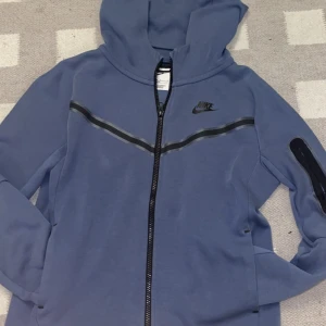 Blå hoodie från Nike - Säljer en snygg blå hoodie från Nike med dragkedja och huva. Den har ett stilrent svart Nike-logotyptryck på bröstet och svarta detaljer längs dragkedjan. Perfekt för en avslappnad stil.