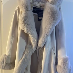 Vit pälsjacka från Caroline - Säljer en elegant vit pälsjacka från Caroline med fluffig fuskpäls på krage och ärmslut. Jackan har en stilren design med bälte i midjan och dragkedja framtill. Perfekt för kyliga dagar när du vill känna dig extra lyxig. Ny med pris lap 