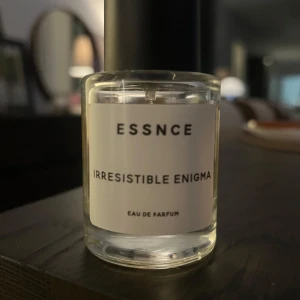 Essnce Irresistible Enigma Eau de Parfum - Upptäck Essnce Irresistible Enigma Eau de Parfum i en elegant glasflaska med svart lock. Denna doft är både mystisk och lockande, perfekt för den som vill göra ett intryck. Flaskan har en stilren design med en vit etikett och svart text.