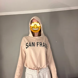 Beige hoodie med tryck - Säljer en beige hoodie med texten 'San Fran' tryckt på framsidan. Hoodien har två snören ifall man vill att tröjan ska sitta tajtare nertill i och en bekväm luva/huva. Perfekt för en avslappnad stil.