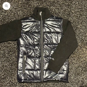 Cardigan med stickade ärmar - Snygg svart cardigan med glansig finish och stickade ärmar från All Cap One. Jackan har en dragkedja framtill. Perfekt för kyligare dagar och nu till våren! Helt oanvänd, säljer pga för liten denna cardigan är helt slutsåld!!!