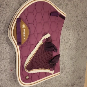 Lila schabrak och huva från Trinity - Säljer ett snyggt lila schabrak med matchande huva från Trinity. Schabraket har ett quiltat mönster och detaljer i beige. Huvan är dekorerad med ett broderat emblem. Perfekt för ridning och att ge din häst en stilren look.