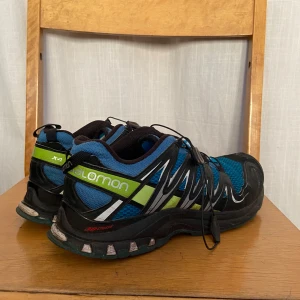 Blå och svarta löparskor från Salomon - Säljer ett par Salomon löparskor i blått och svart med gröna detaljer. Skorna har snörning och en robust sula, perfekt för terränglöpning. De är designade för att ge bra grepp och stabilitet.