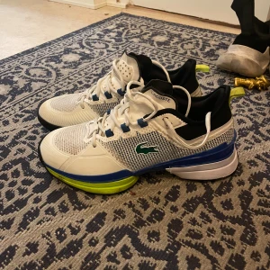 Vita sneakers från Lacoste - Snygga vita sneakers från Lacoste med blå och gröna detaljer. Skorna har snörning och en sportig design med Lacoste-loggan på sidan. Perfekta för en aktiv livsstil.