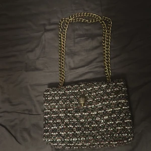 Mönstrad axelväska från Kurt Geiger - Snygg axelväska från Kurt Geiger med ett unikt mönster i svart, vitt och rött. Väskan har en elegant kedjerem och ett dekorativt spänne framtill. Perfekt för att ge din outfit en stilfull touch. Ska dock nämna att kedjan har börjat färga av sig lite om man har exempel en vit T-shirt som går bort i tvätten men det kan fixas genom att exempelvis måla me ett genomskinligt nagellack 