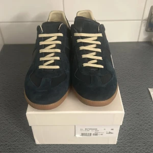 Mörkblå sneakers med beige snörning - Snygga mörkblå sneakers från Maison Margiela med beige snörning och ljusbrun sula. Skorna har en stilren design med en kombination av mocka och syntetmaterial. Perfekta för en trendig och avslappnad stil. Säljer de för att de är för små så har ingen användning av de.