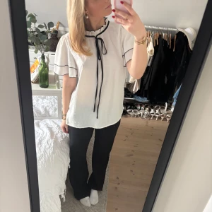 Vit blus med svarta detaljer - Säljer en söt vit blus med svarta kantdetaljer och knytband vid halsen. Blusen har korta ärmar med volang och prickigt mönster. Perfekt till fest och vardag. Använd fåtal gånger🥰