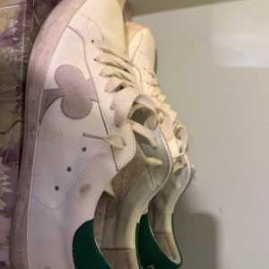 The gallgerist  - Intressekoll! Säljer ett par vita sneakers med gröna detaljer och snörning. Skorna har en unik design med ett klubbmönster på sidan. Perfekta för en avslappnad stil.