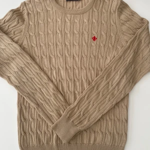 Beige kabelstickad tröja från Morris - Säljer en stilren beige kabelstickad tröja från Morris i 100% merceriserad bomull. Tröjan har ett rött broderat emblem på bröstet och ribbade muddar vid ärmslut och nederkant. Perfekt för en klassisk look!