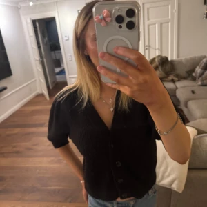 Svart kofta med knappar - Säljer en stilren svart kofta med knappar framtill. Koftan har korta ärmar och en v-ringad design, perfekt för en avslappnad look. Passar bra till jeans eller kjol.