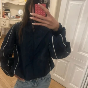 Svart bomberjacka med vita detaljer - Snygg svart bomberjacka med vita kontrastlinjer. Jackan har en loose passform och långa ärmar. Perfekt för en cool och avslappnad stil.