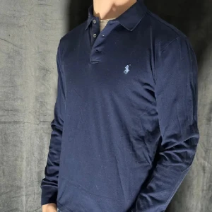  Ralph Lauren Tröja - Säljer en stilren mörkblå pikétröja från Ralph Lauren. Tröjan har en klassisk krage med knappar och det ikoniska logotypbroderiet på bröstet. Perfekt för en avslappnad men ändå elegant look.