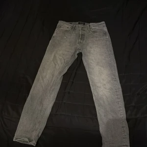 Grå jeans från Jack & Jones - Snygga grå jeans från Jack & Jones med en tapered passform. De har en klassisk femficksdesign och en liten sliten detalj på framsidan för en cool look. Perfekta för en avslappnad stil.