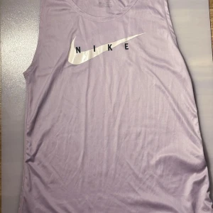 Nine swoosh träningslinne  - Snygg ljus lila/rosa ärmlös topp från Nike med deras ikoniska logga i vitt på framsidan. Perfekt för träning med Dri-FIT material som håller dig sval och torr. Enkel och stilren design som passar till många olika outfits.