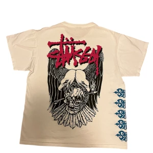 Stüssy Tshirt - Säljer en cool vit t-shirt från Stüssy med ett stort grafiskt tryck i svart och rött på baksidan. Framsidan har ett mindre tryck på bröstet och blå detaljer längs sidan. Den är nästan helt ny och äkta har kvitto