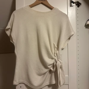 Vit topp från Zara - Säljer en stilren vit topp från Zara med korta ärmar och en snygg knytdetalj på sidan. Perfekt för en avslappnad men chic look. Tillverkad i ett mjukt material som känns skönt mot huden.