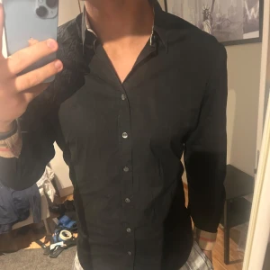 Burberry skjorta - Snygg svart skjorta från Burberry med klassisk krage och knappar framtill. Skjortan har en stilren design och är perfekt för en elegant look. Den har långa ärmar och en diskret randig insida vid kragen.