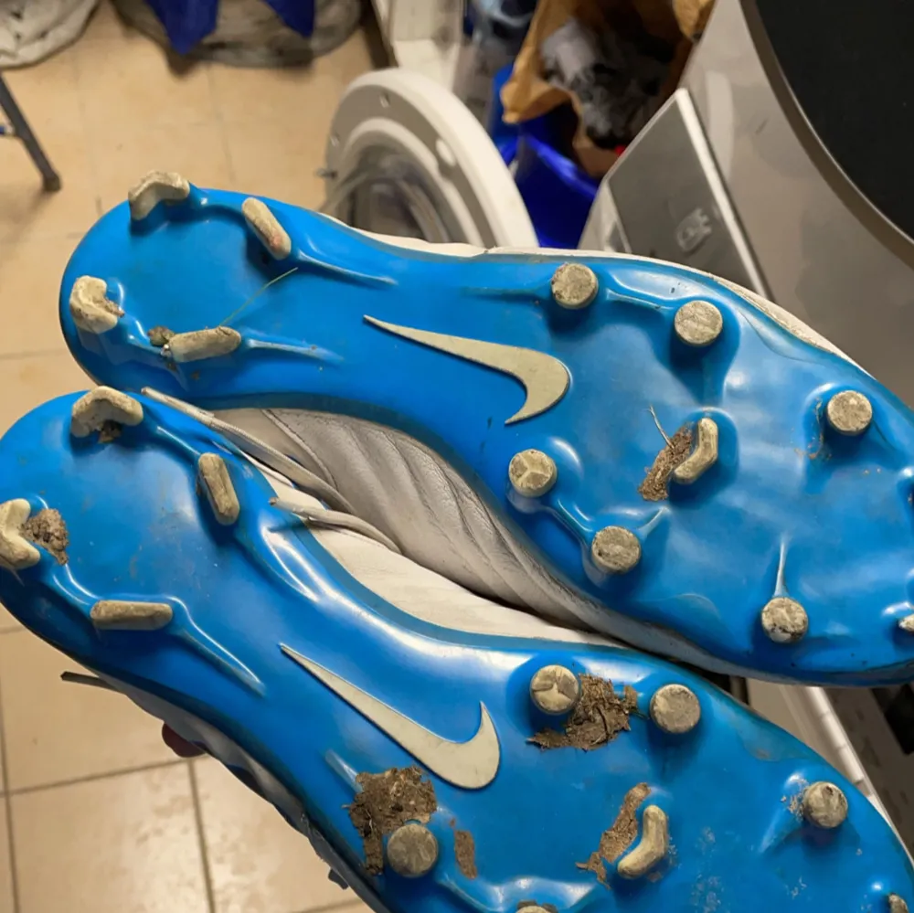 Säljer ett par vita Nike fotbollsskor med blå detaljer och snörning. Skorna har en blå sula med dobbar, perfekt för gräsplaner. Modellen är Nike Tiempo, känd för sin komfort och hållbarhet.. Kengät.