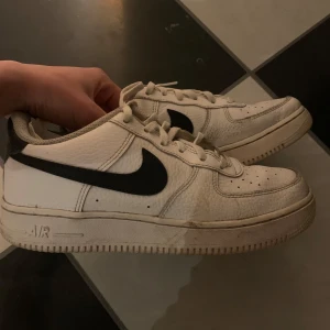 Nike Air Force i vitt och svart - Säljer ett par klassiska Nike Air Force sneakers i vitt med svart swoosh. Skorna har snörning och är tillverkade i skinn. Perfekta för en stilren look.