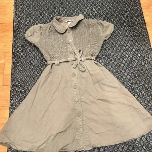 Grön klänning med knappar - Charmig grön klänning med korta ärmar och knappar framtill. Den har en söt krage och ett knytband i midjan för en fin passform. Perfekt för en avslappnad stil.