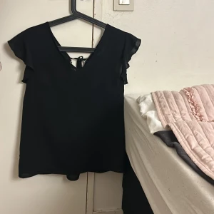 Svart blus med spetsdetaljer - Elegant svart blus med v-ringning och spetsdetaljer. Blusen har korta ärmar med volang och knytning framtill för en stilfull look.