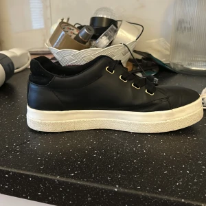 Svarta sneakers från Gant - Snygga svarta sneakers från Gant med vit sula och gulddetaljer. Skorna har snörning och en diskret logga på hälen. Perfekta för en stilren look.